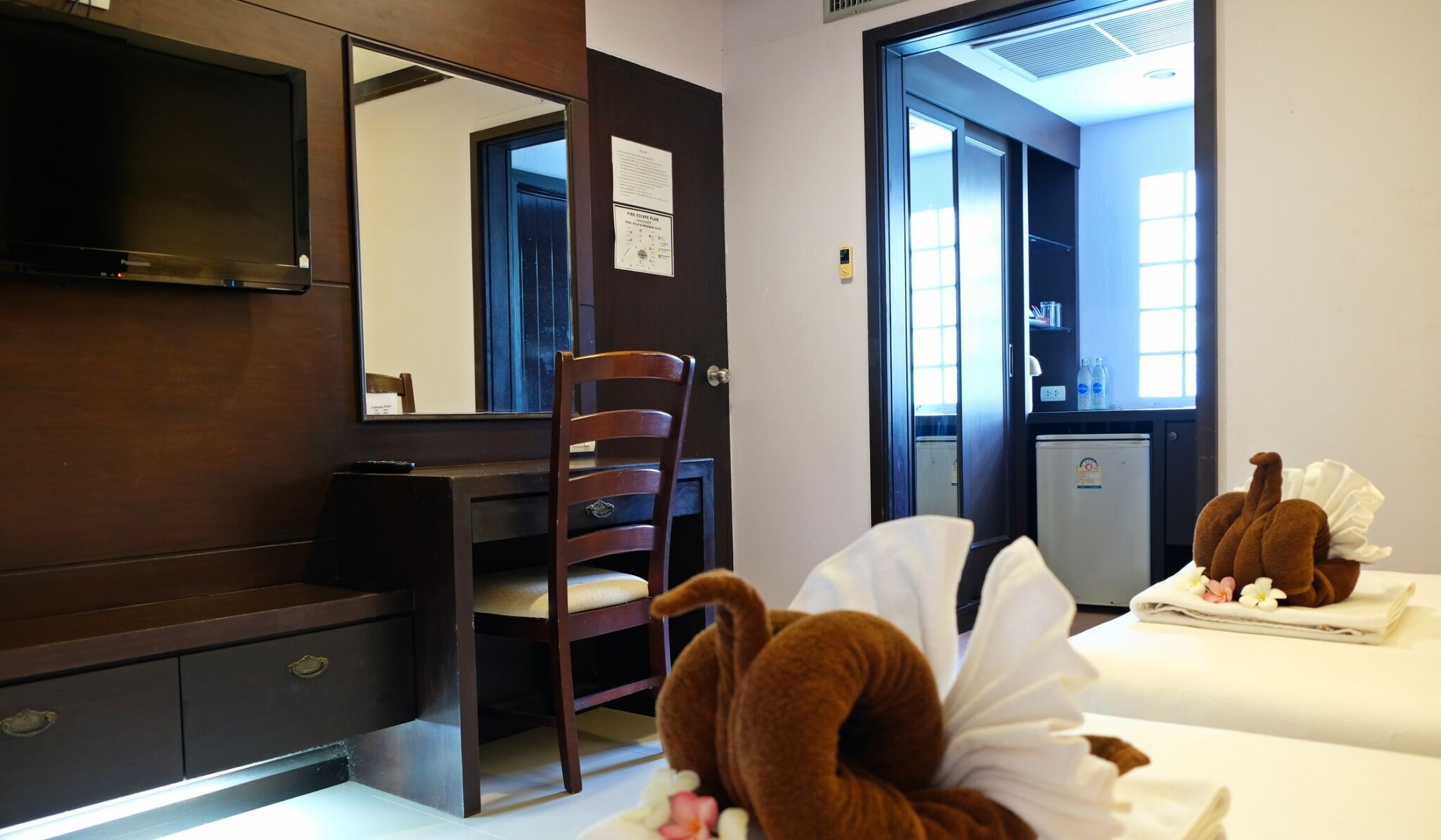 A hotel room with a wooden desk, a chair, a bed with a plush toy, a small refrigerator, and a window with a view.