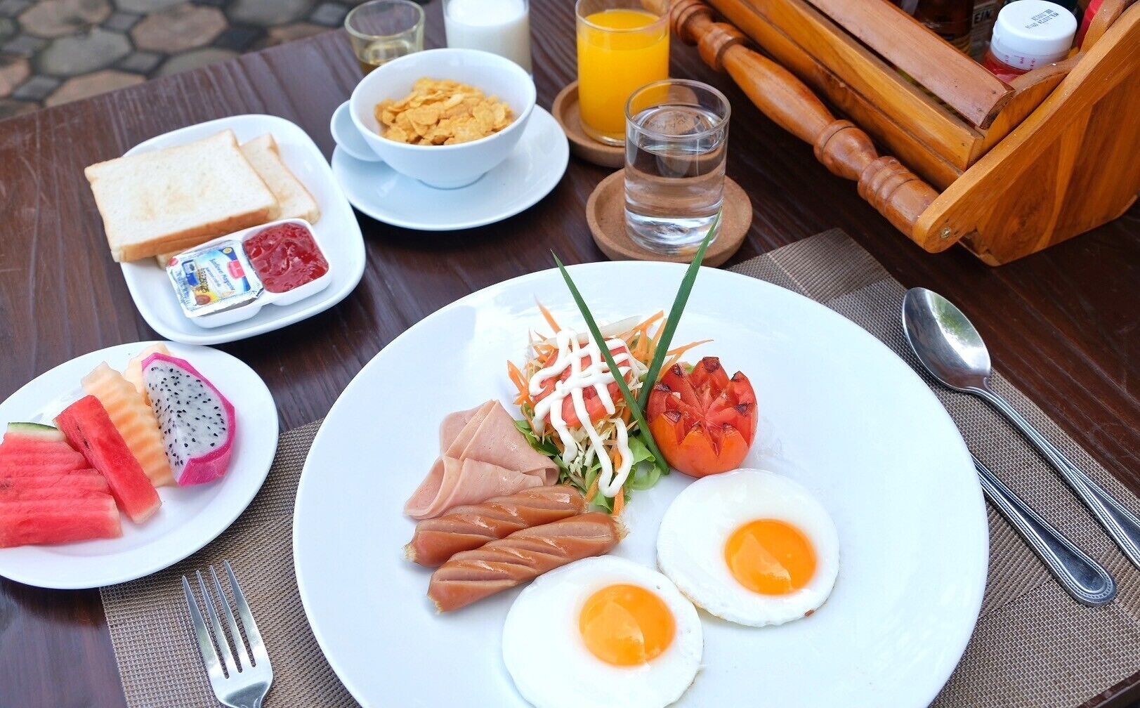 A breakfast plate with eggs, meat, and vegetables, accompanied by toast, fruit, and a bowl of cereal.