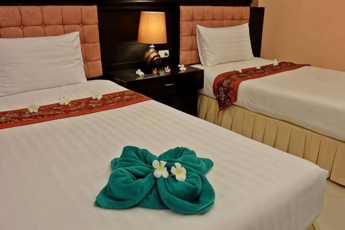 Duas camas com lençóis brancos, uma cabeceira, uma mesinha de cabeceira com abajur e um objeto decorativo vermelho sobre a cama.