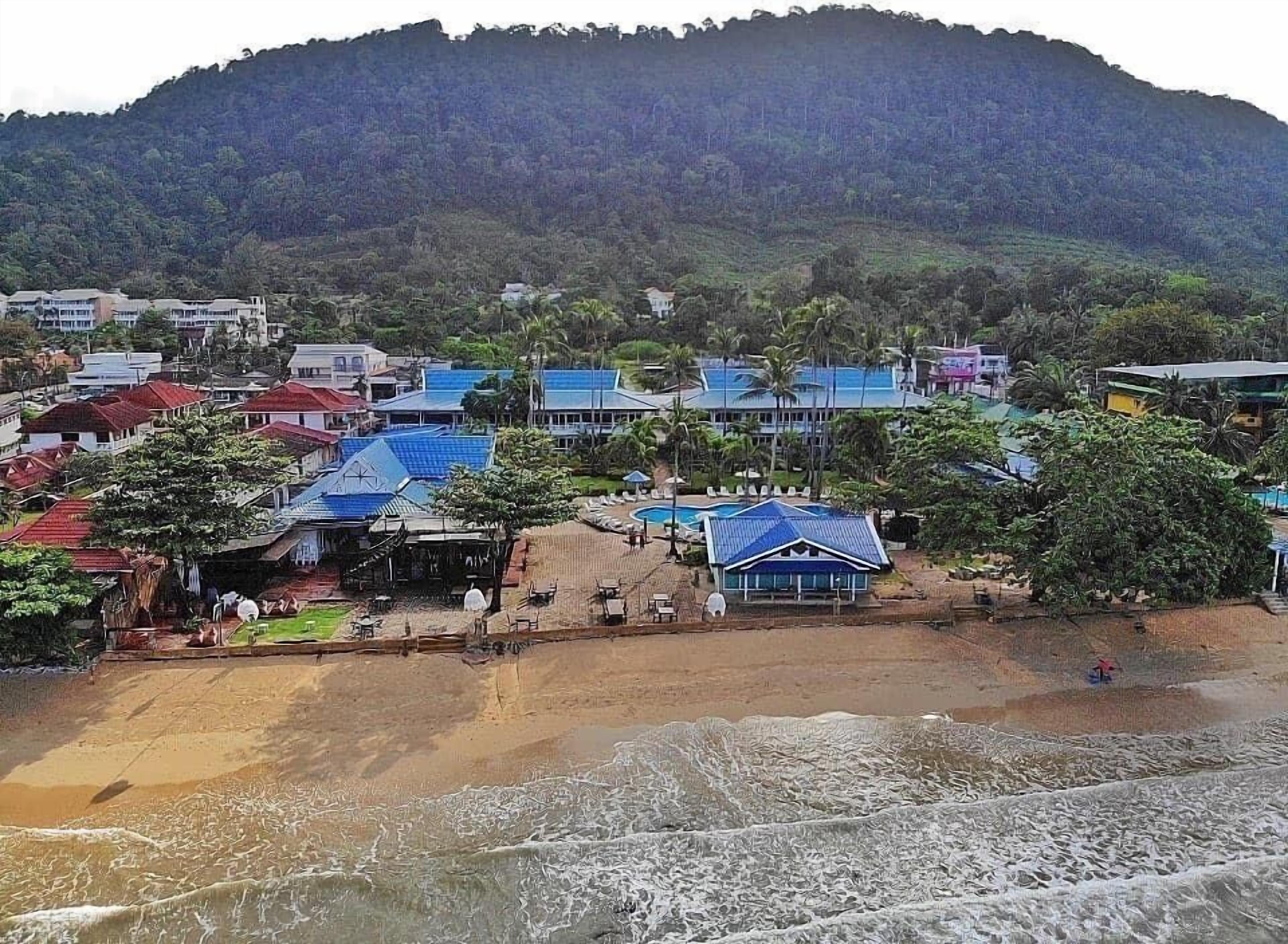Un complejo turístico frente al mar con acceso a la playa, piscina y vistas a una colina boscosa.