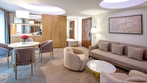 Quarto de hotel moderno com uma mesa de jantar redonda, sofás bege, uma mesa de centro e uma grande pintura abstrata na parede.