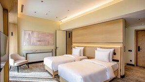 Quarto de hotel com duas camas, uma cadeira, um quadro abstrato grande e um radiador.