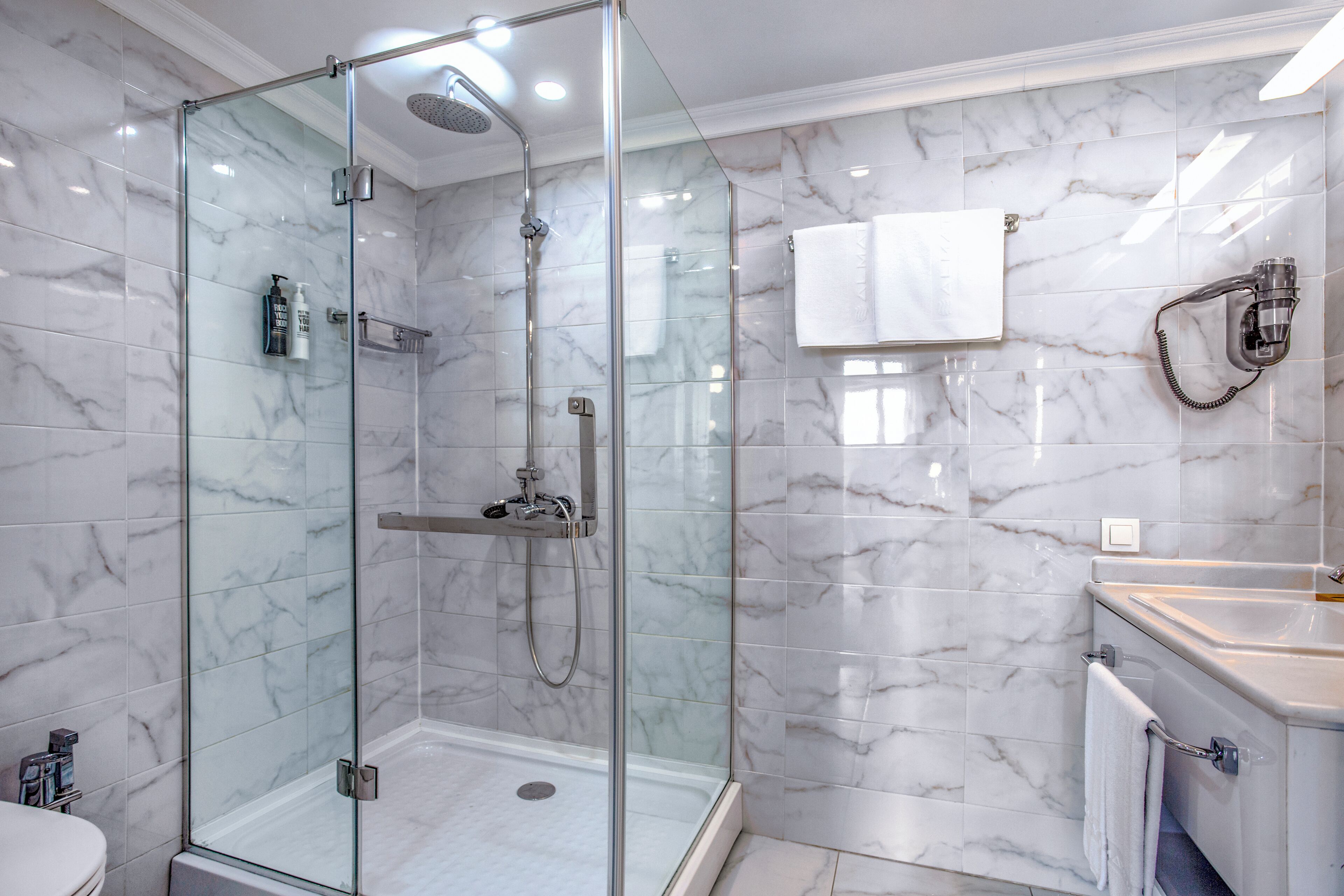 Un baño moderno con ducha de vidrio, azulejos de mármol, un lavamanos blanco y un perchero.