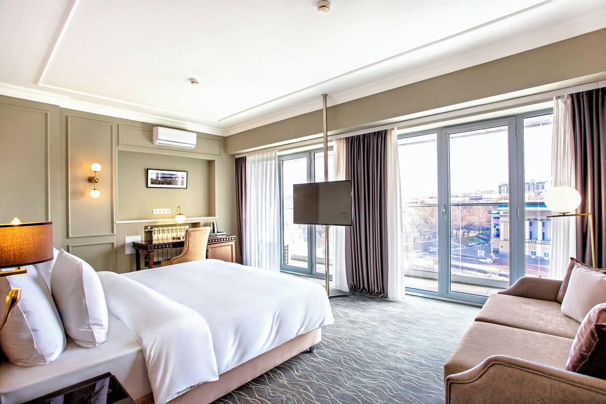 A hotel room with a bed, a sofa, a desk with a lamp, a TV, and large windows with curtains.