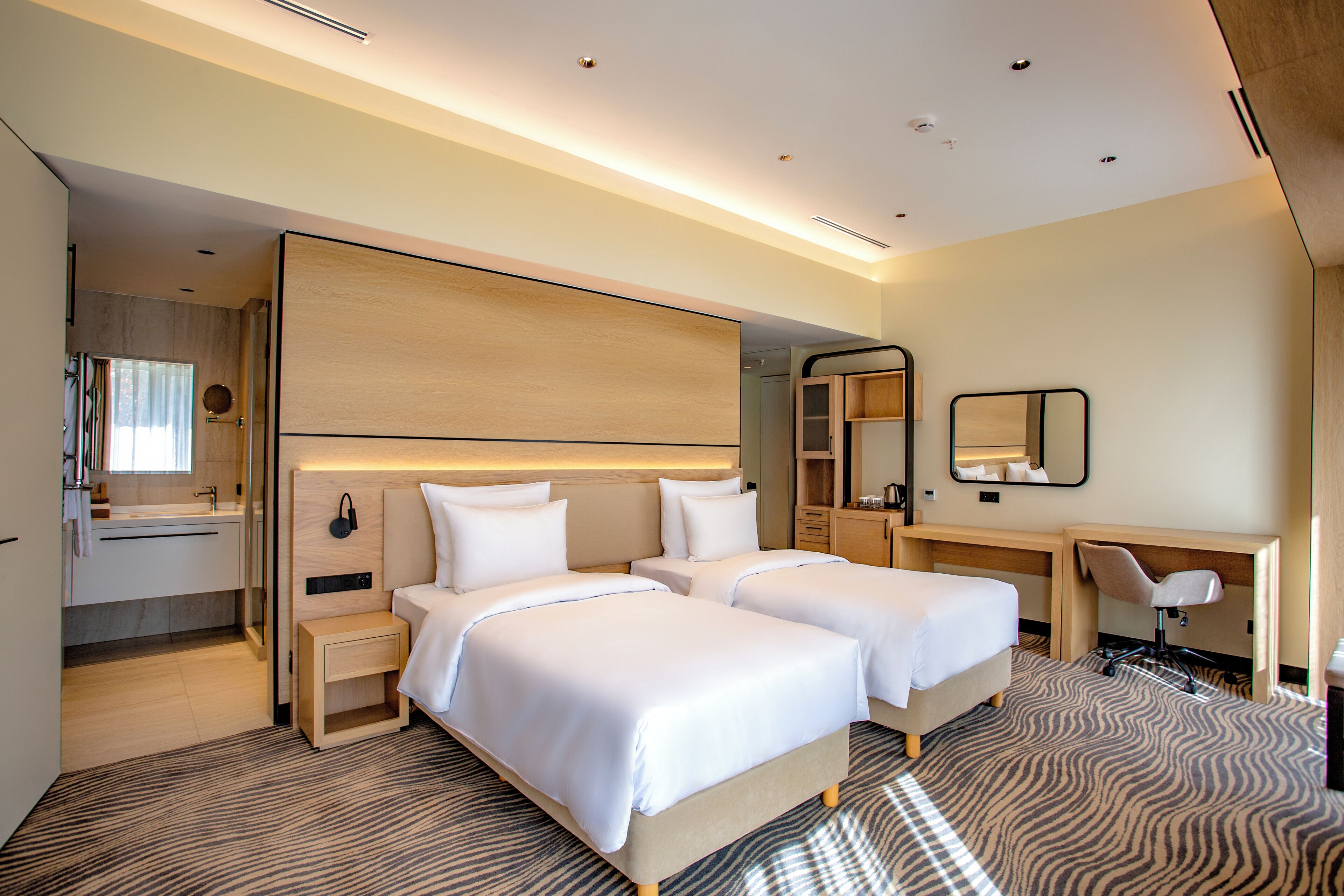 Habitación de hotel con dos camas, un escritorio, una silla, un espejo y un baño visible a través de una puerta abierta.