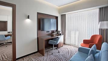 A hotel room with a desk, a television, a sofa, and a large window with curtains.