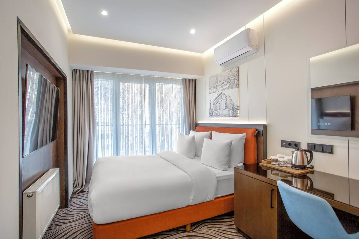 A hotel room with a bed, a desk, a chair, a TV, and a window with curtains.