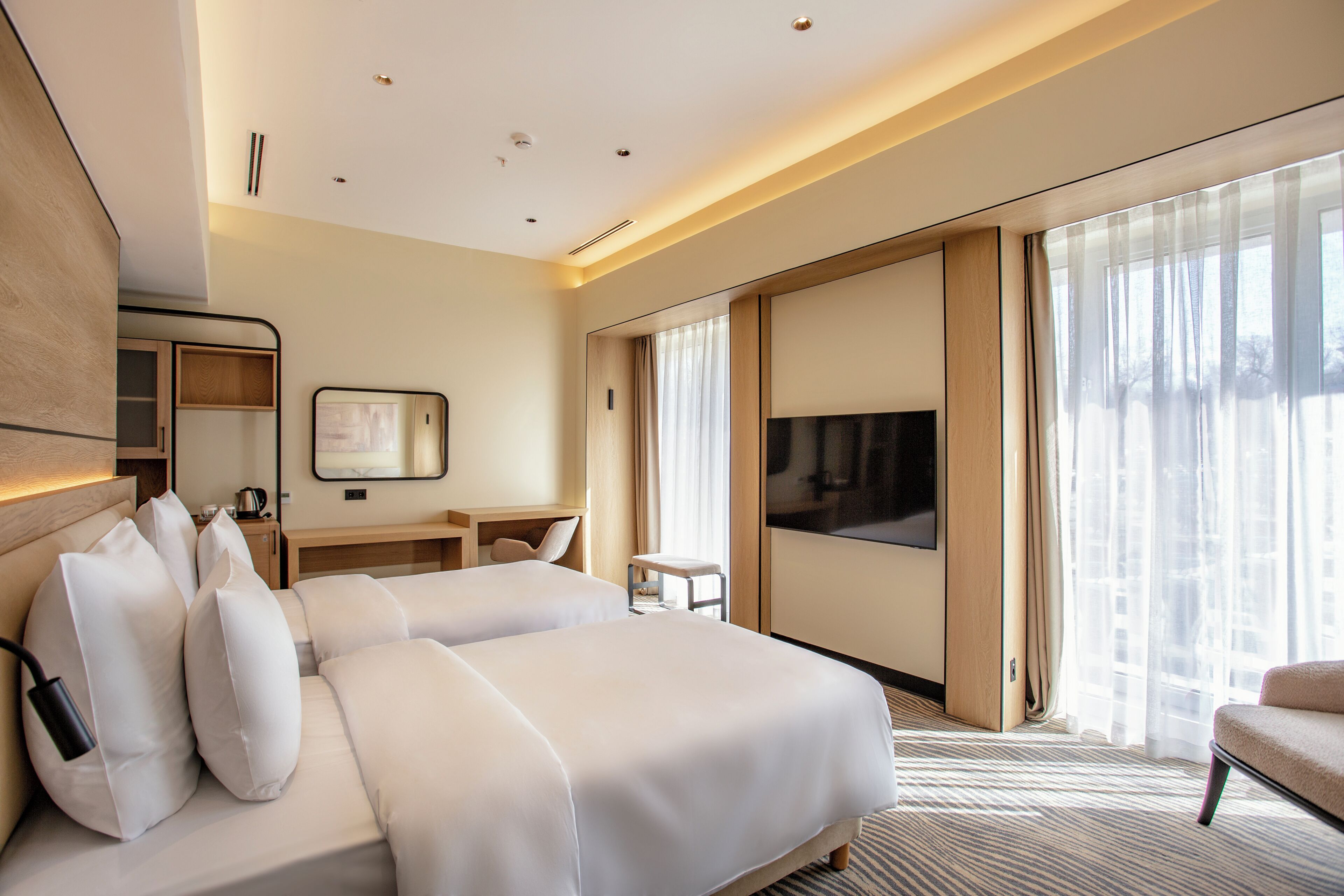 A hotel room with two beds, a desk, a chair, a TV, and large windows.