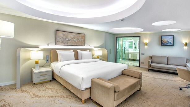 Quarto de hotel moderno com uma cama grande, mesas de cabeceira, um sofá e uma porta de vidro para uma varanda.
