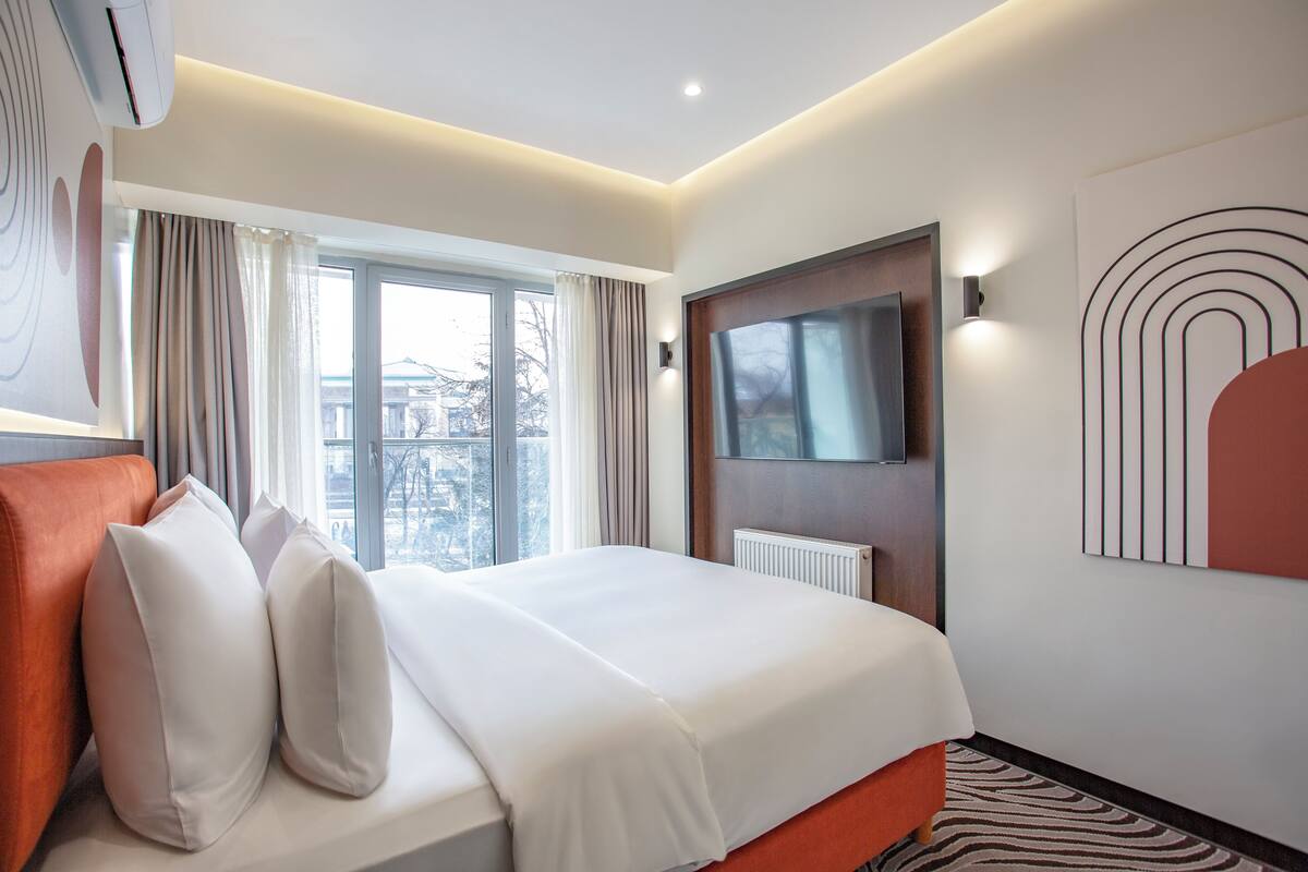 A modern hotel room with a large bed, a television mounted on the wall, and a window with curtains.
