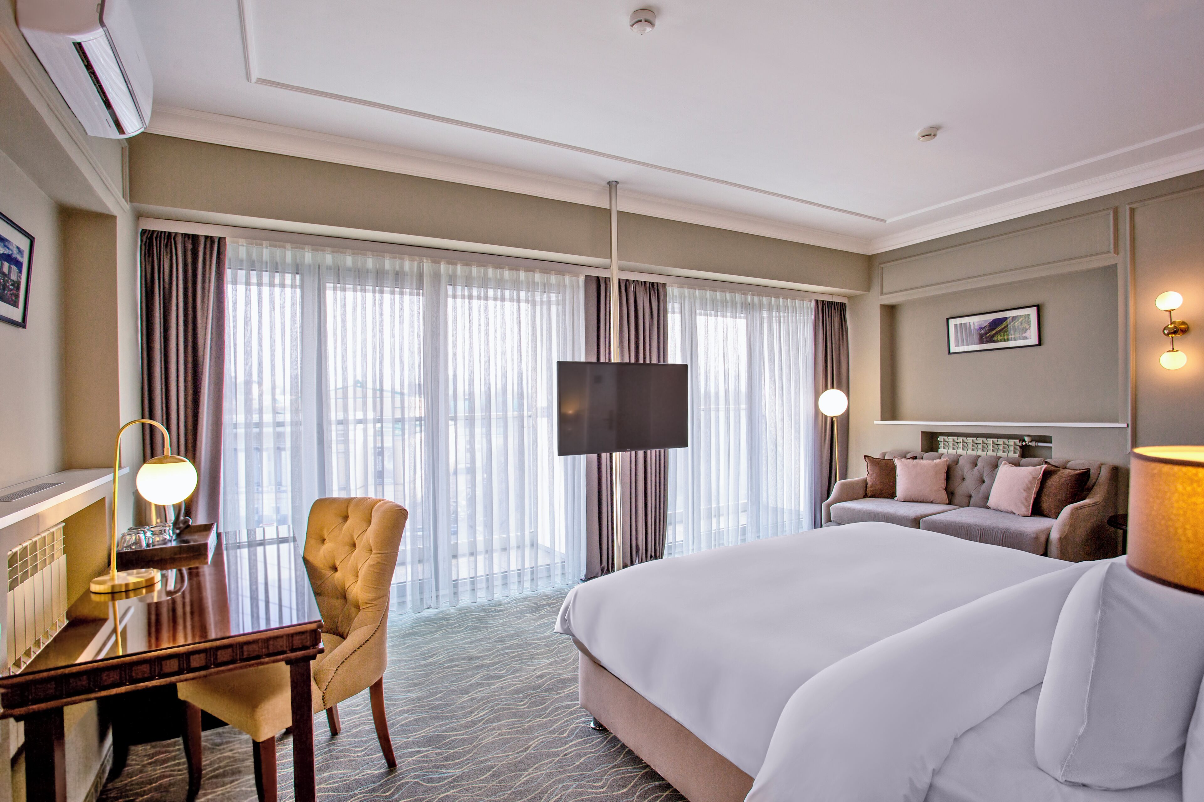 A hotel room with a bed, a desk with a lamp, a chair, a TV, and a sofa.
