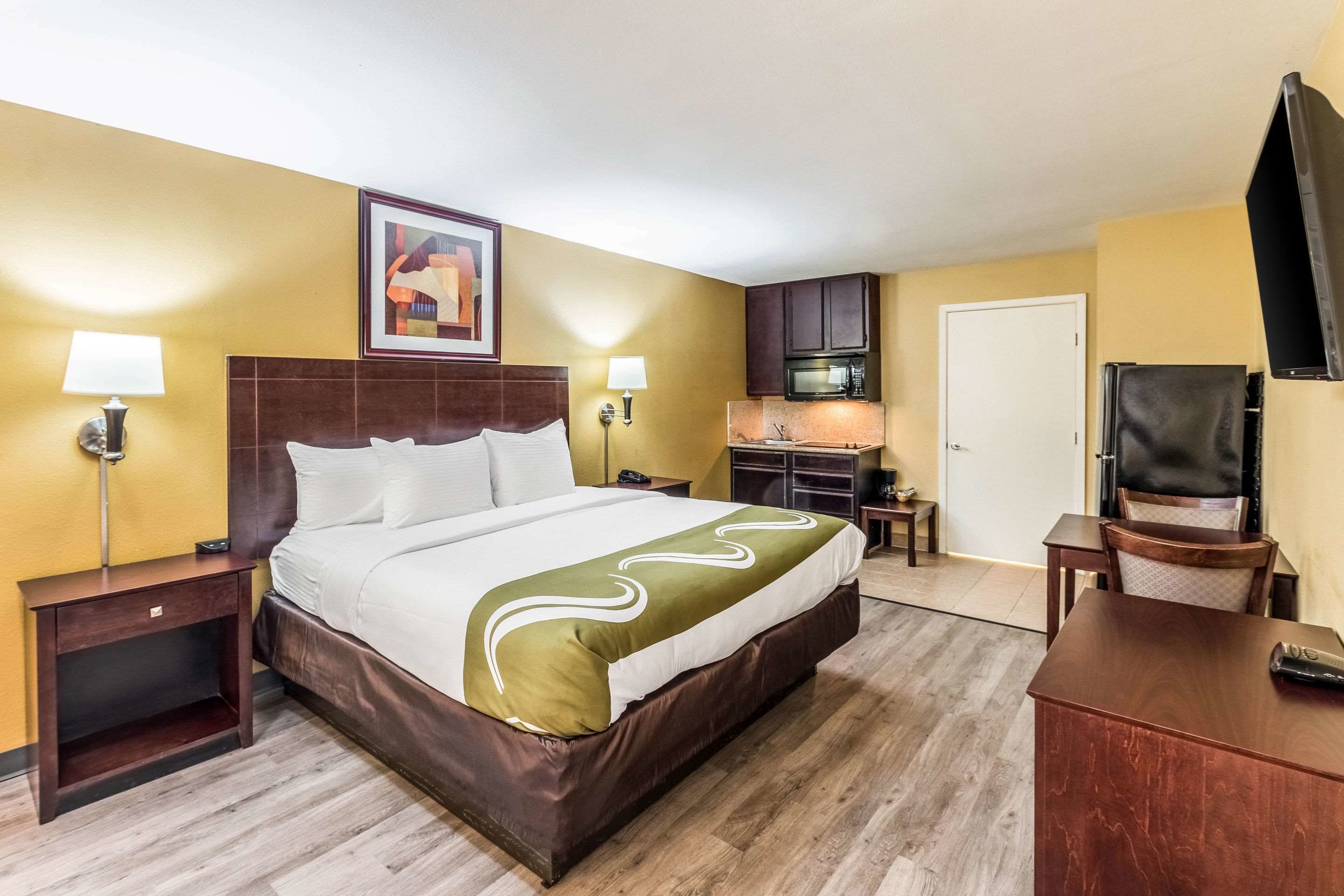 A hotel room with a bed, bedside tables, a desk, a chair, a TV, a microwave, and a door.