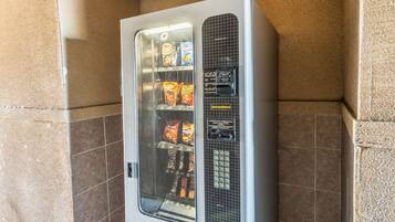 A vending machine with snacks and beverages, located in a corner with tiled walls.