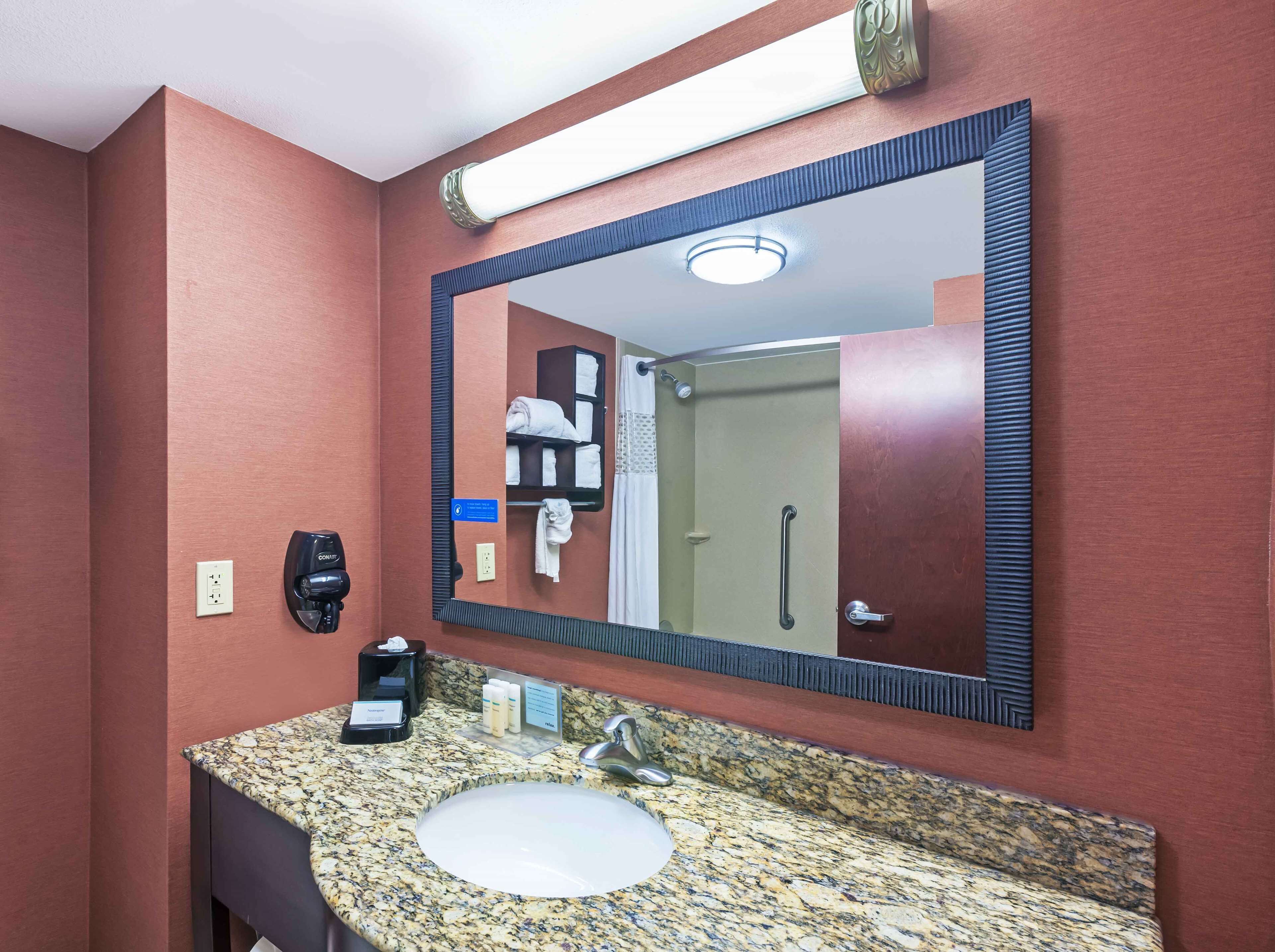 A bathroom with a granite countertop sink, a large mirror, a towel rack, a hand dryer, and a sign indicating amenities.