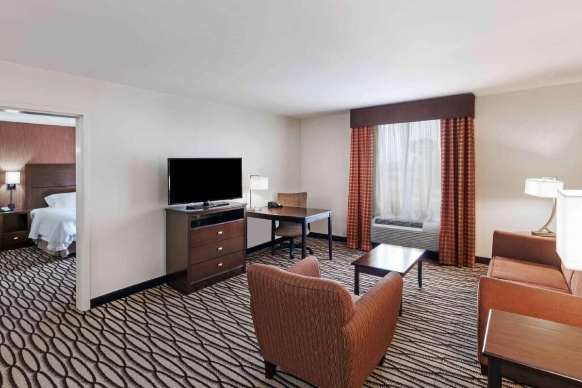 Habitación de hotel con cama, televisión, escritorio, silla, sofá y ventana con cortinas.