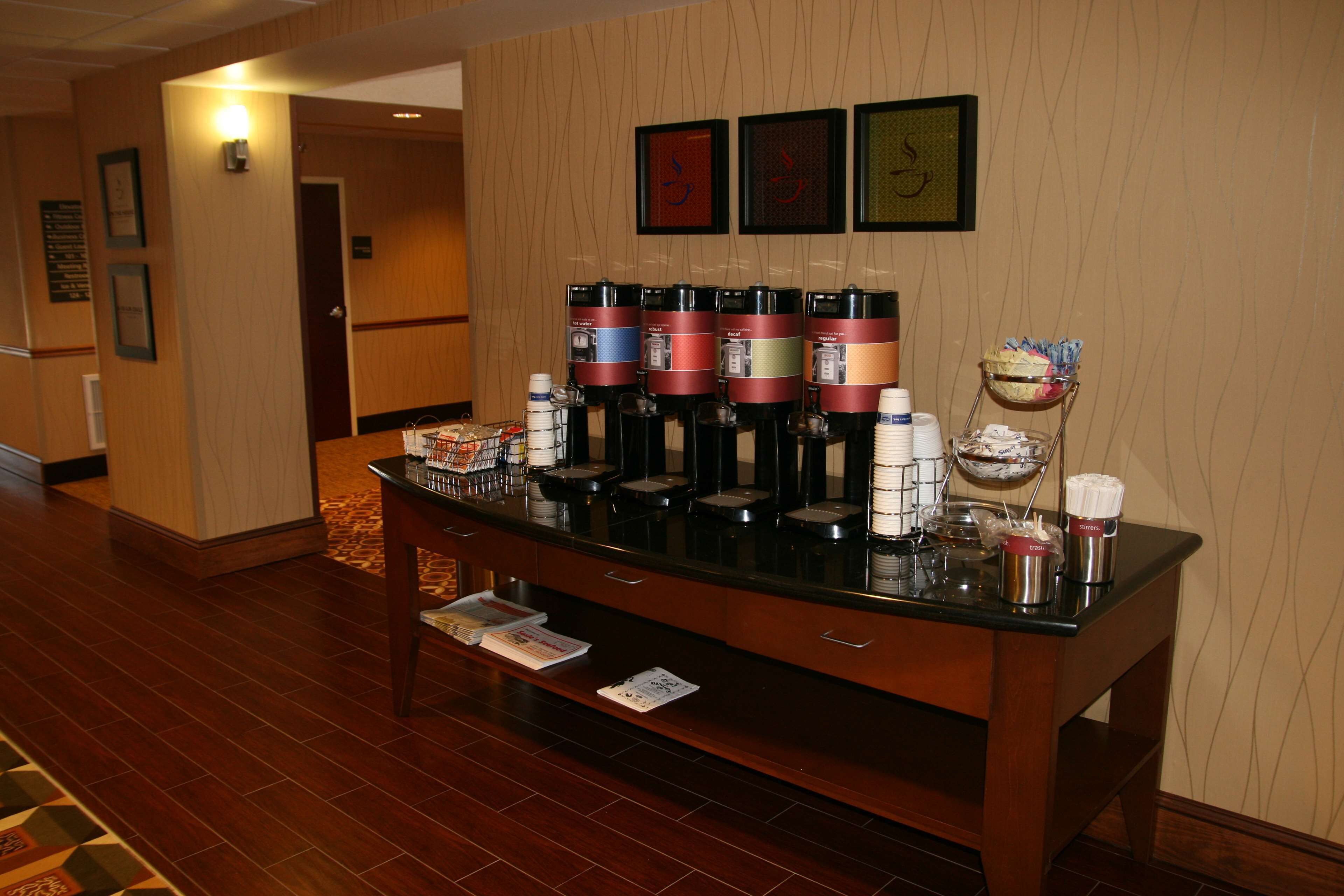 A hotel reception area with a coffee station featuring multiple coffee dispensers, a fruit bowl, and a small refrigerator.