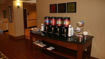 A hotel reception area with a coffee station featuring multiple coffee dispensers, a fruit bowl, and a small refrigerator.