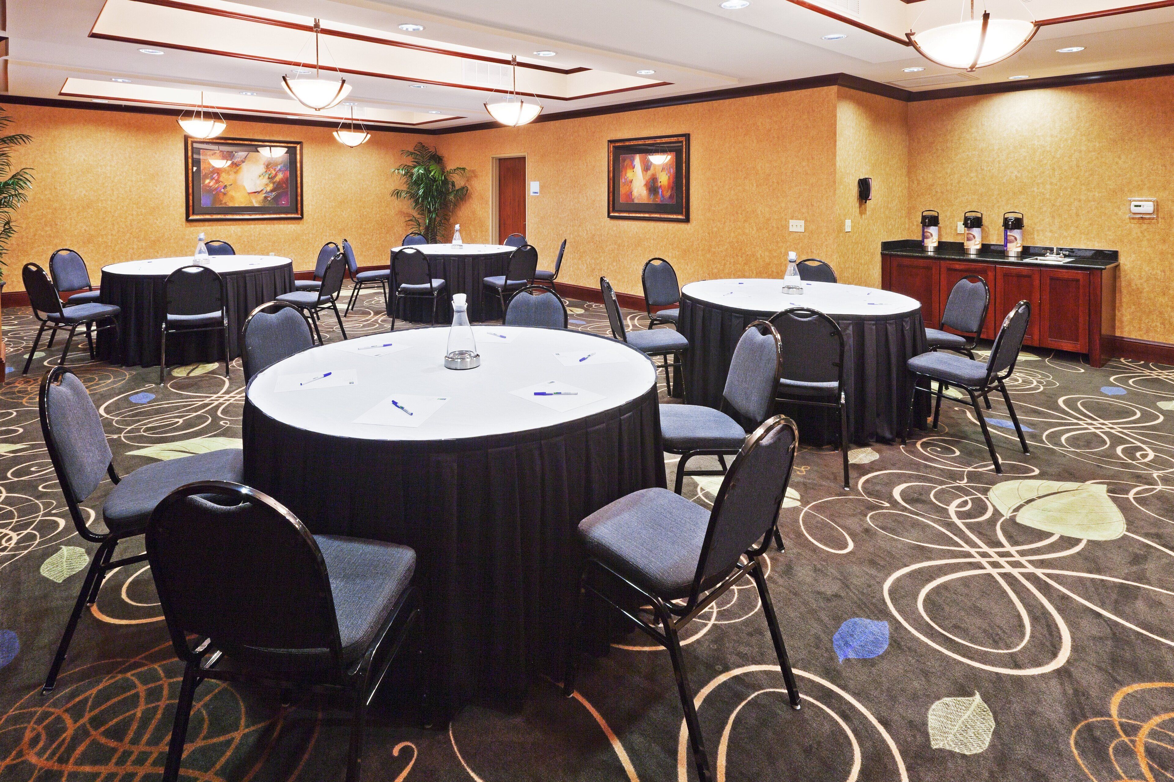 Sala de conferência com mesas e cadeiras redondas, bebedouro e espaço para café.