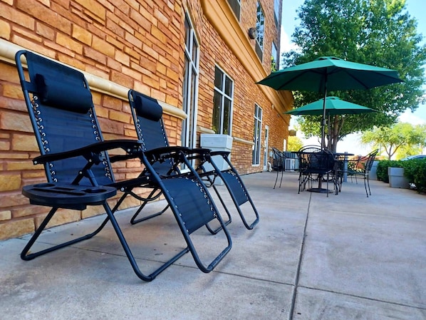 Espace extérieur aménagé avec des chaises et une table, un parasol vert et, en arrière-plan, un bâtiment en briques.