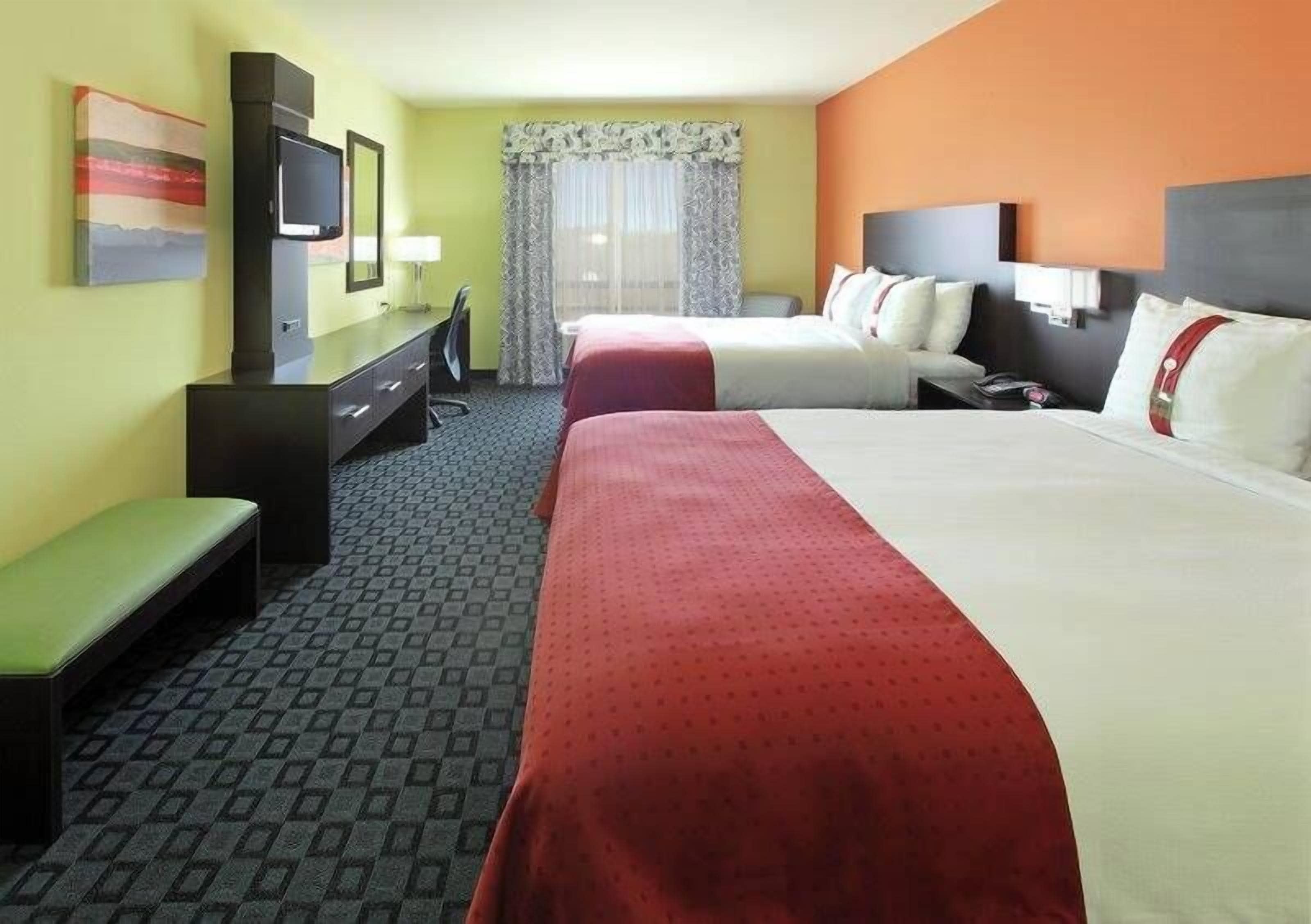 A hotel room with two beds, a desk with a television, a bench, and a window with curtains.