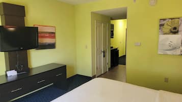 A hotel room with a bed, a TV on a stand, a desk with drawers, a door leading to another room, and a wall with a flower painting.