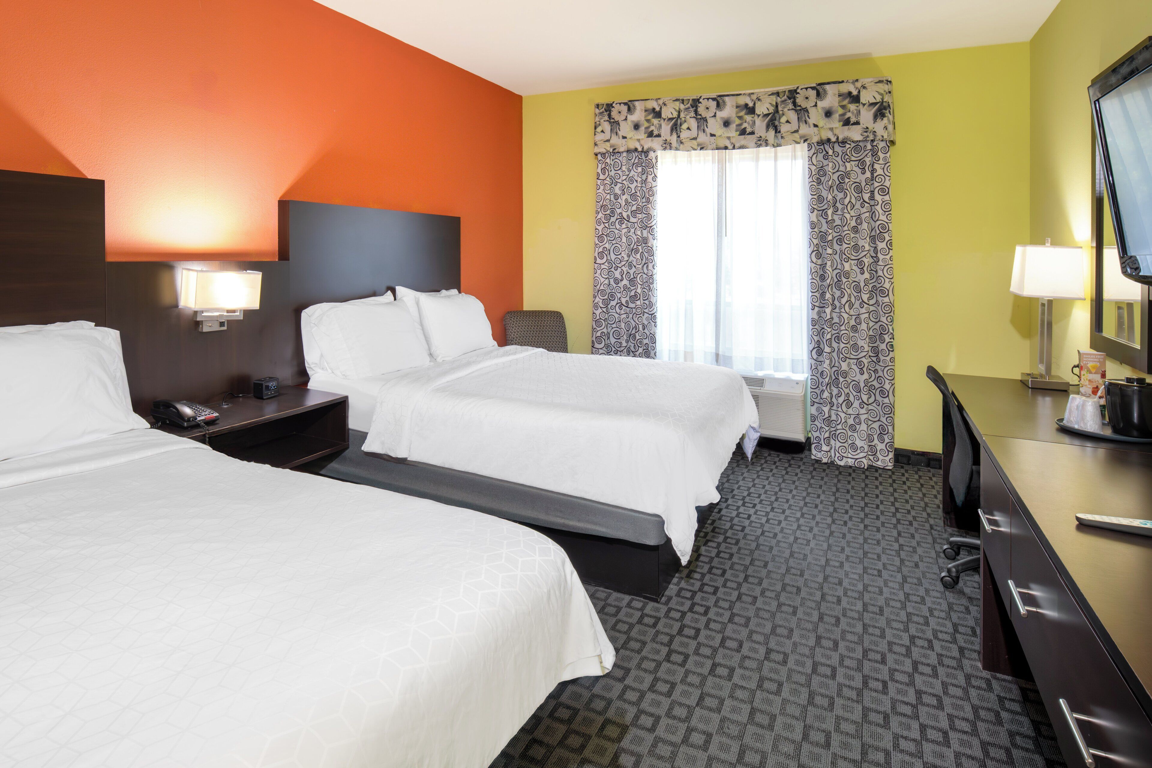 A hotel room with two beds, a desk with a television, and a window with patterned curtains.