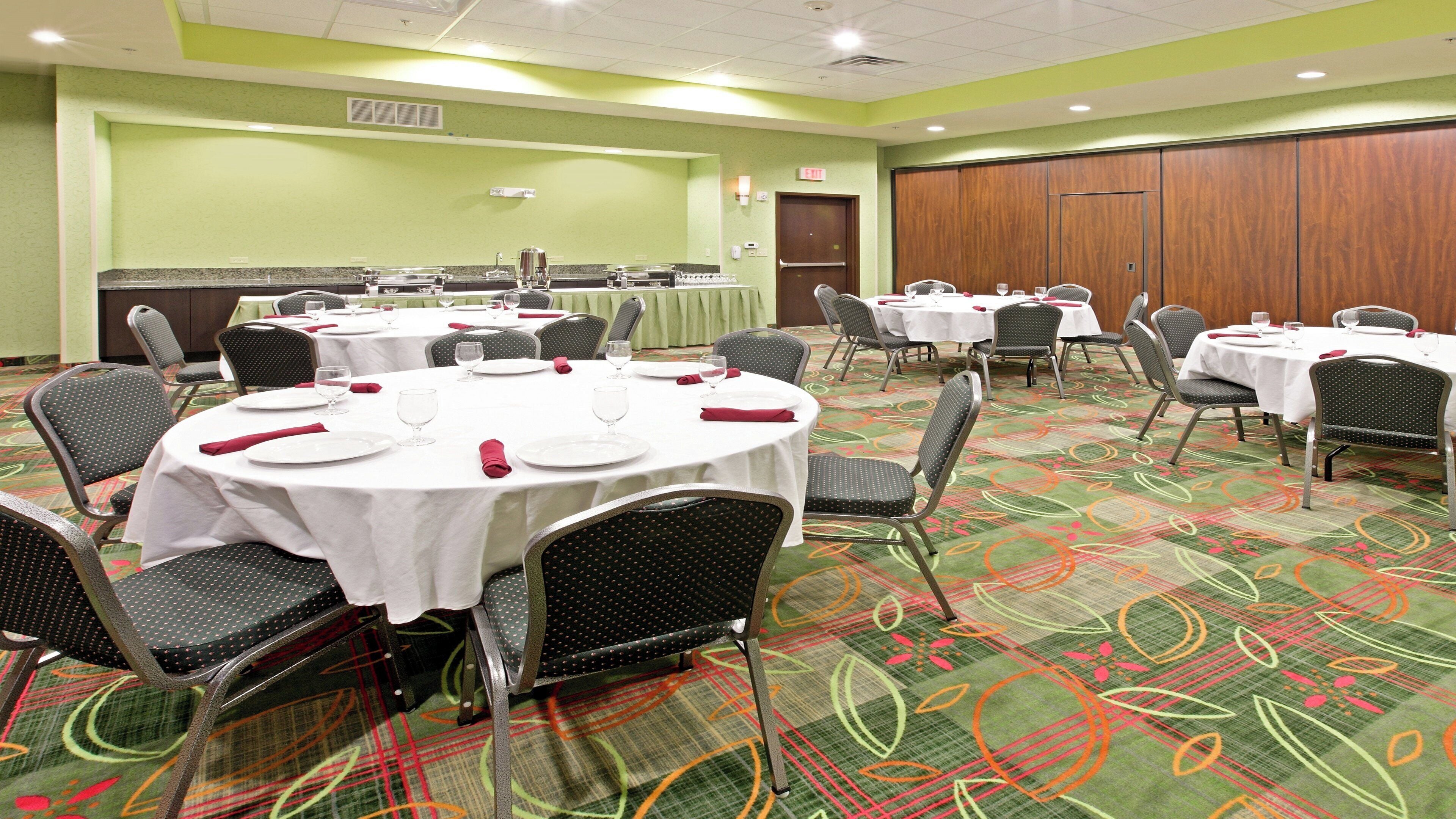 A conference room with multiple round tables, each set with white tablecloths, plates, and red napkins. The room has a green carpet with a floral pattern and is equipped with chairs and a large wooden door.