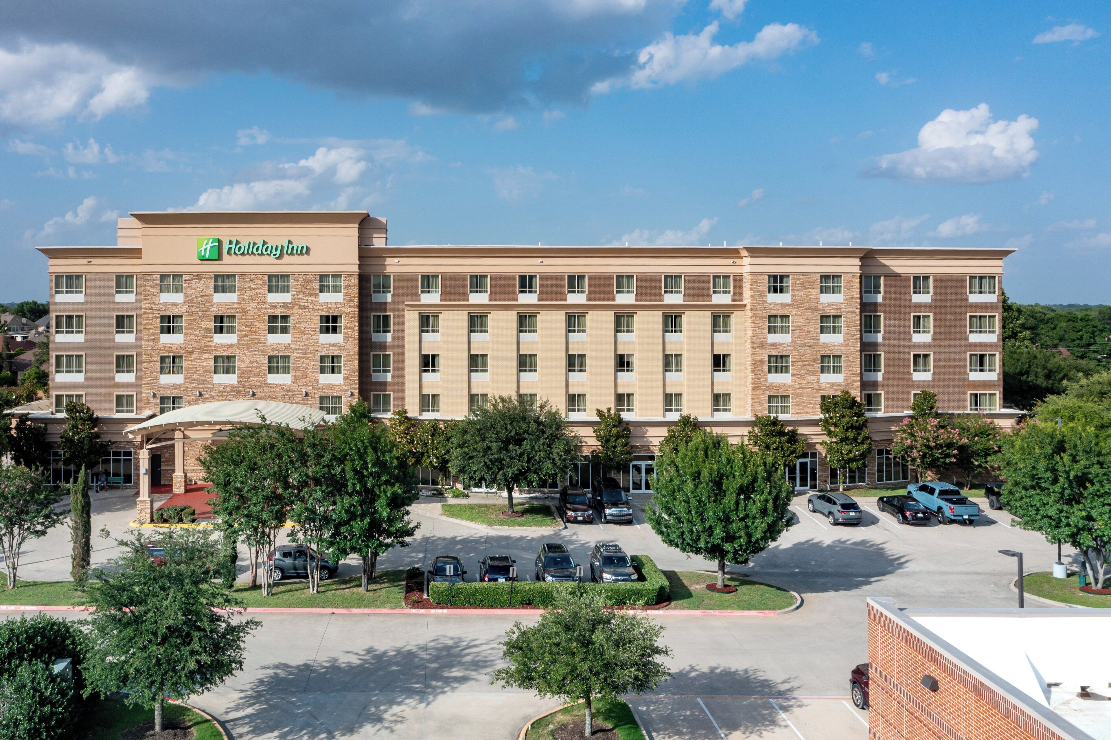 A Holiday Inn hotel with a brick facade, multiple floors, and a parking lot with several cars.