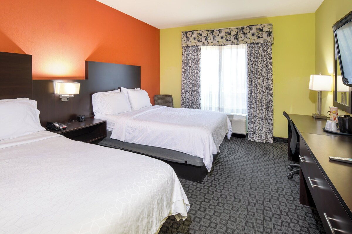 A hotel room with two beds, a desk, a chair, a TV, and a window with patterned curtains.