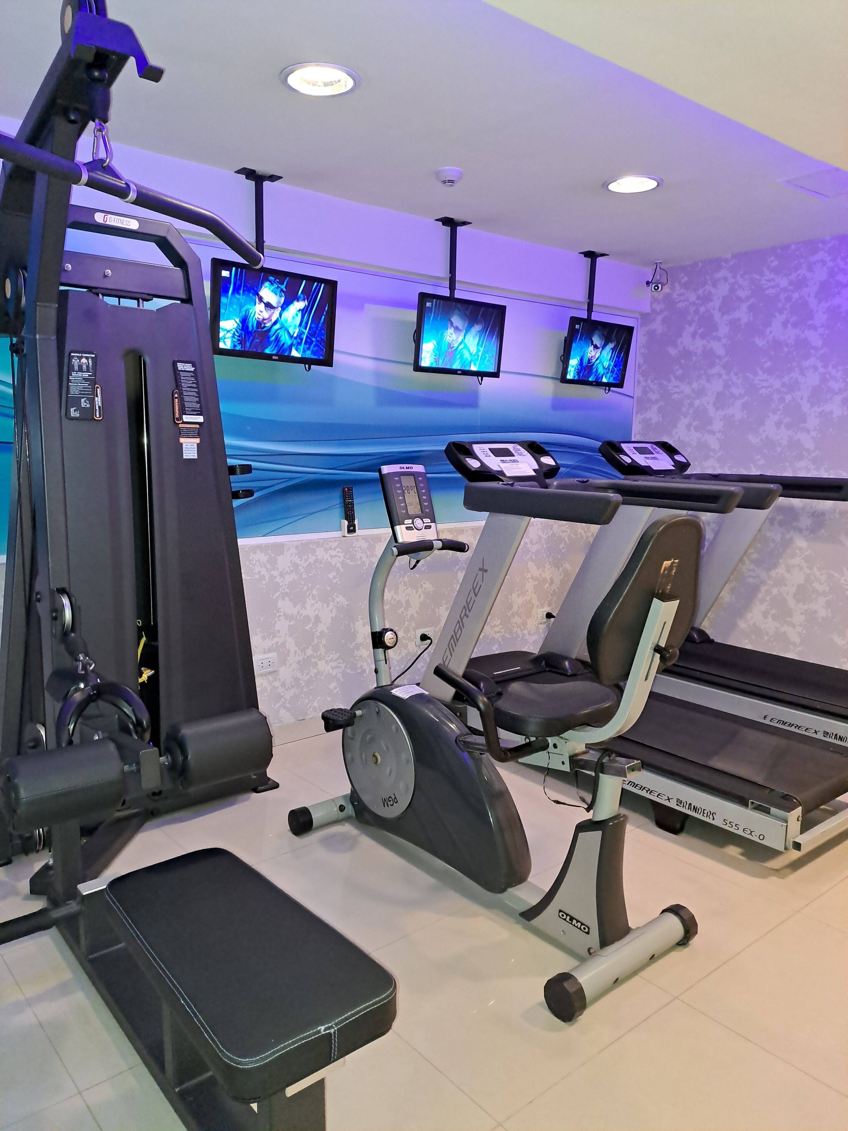 A gym with various exercise equipment, including a treadmill, elliptical, and weight machines. There are three monitors on the wall displaying workout videos.