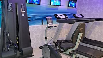 A gym with various exercise equipment, including a treadmill, elliptical, and weight machines. There are three monitors on the wall displaying workout videos.