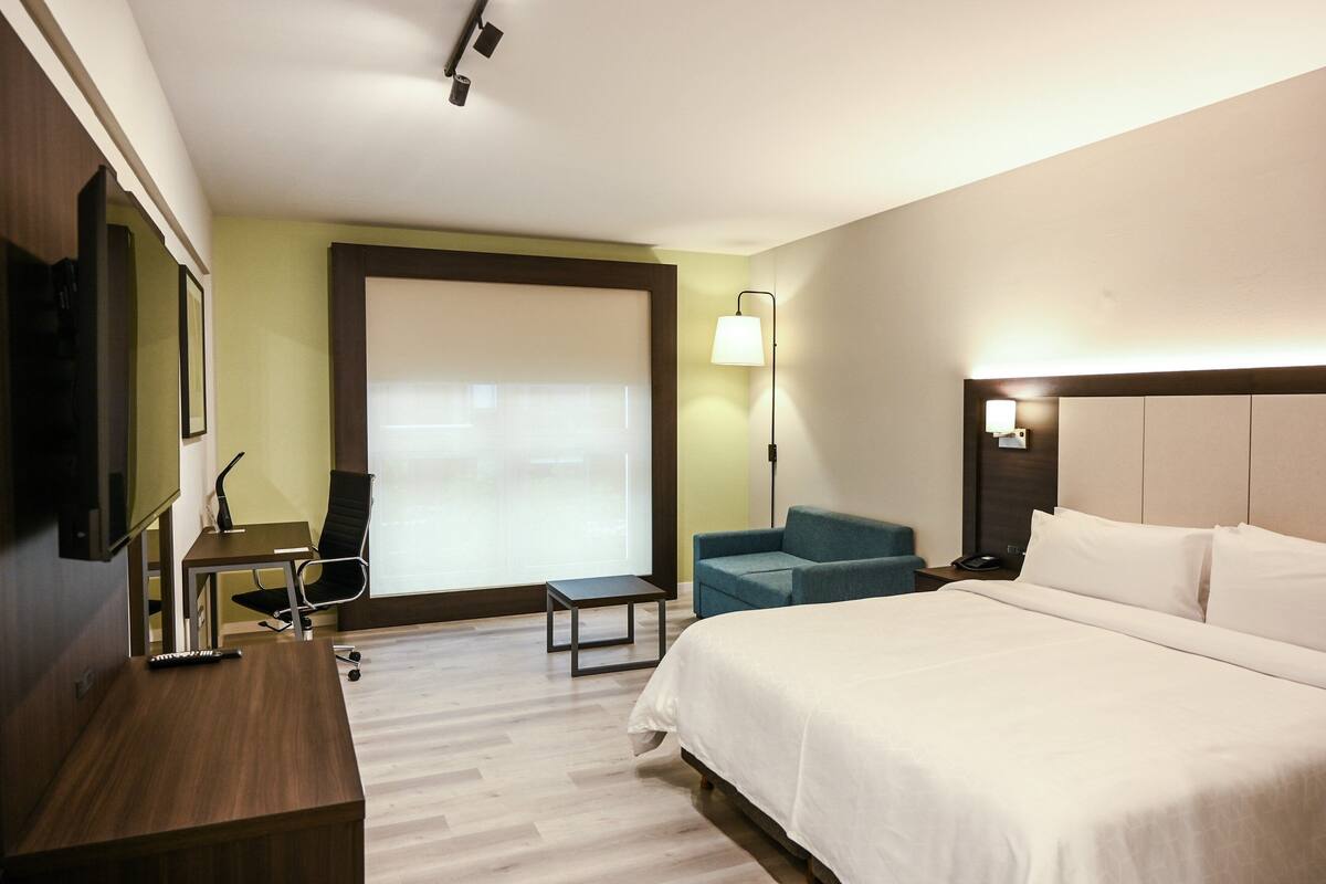 A hotel room with a large bed, a desk, a chair, a sofa, a TV, and a lamp.
