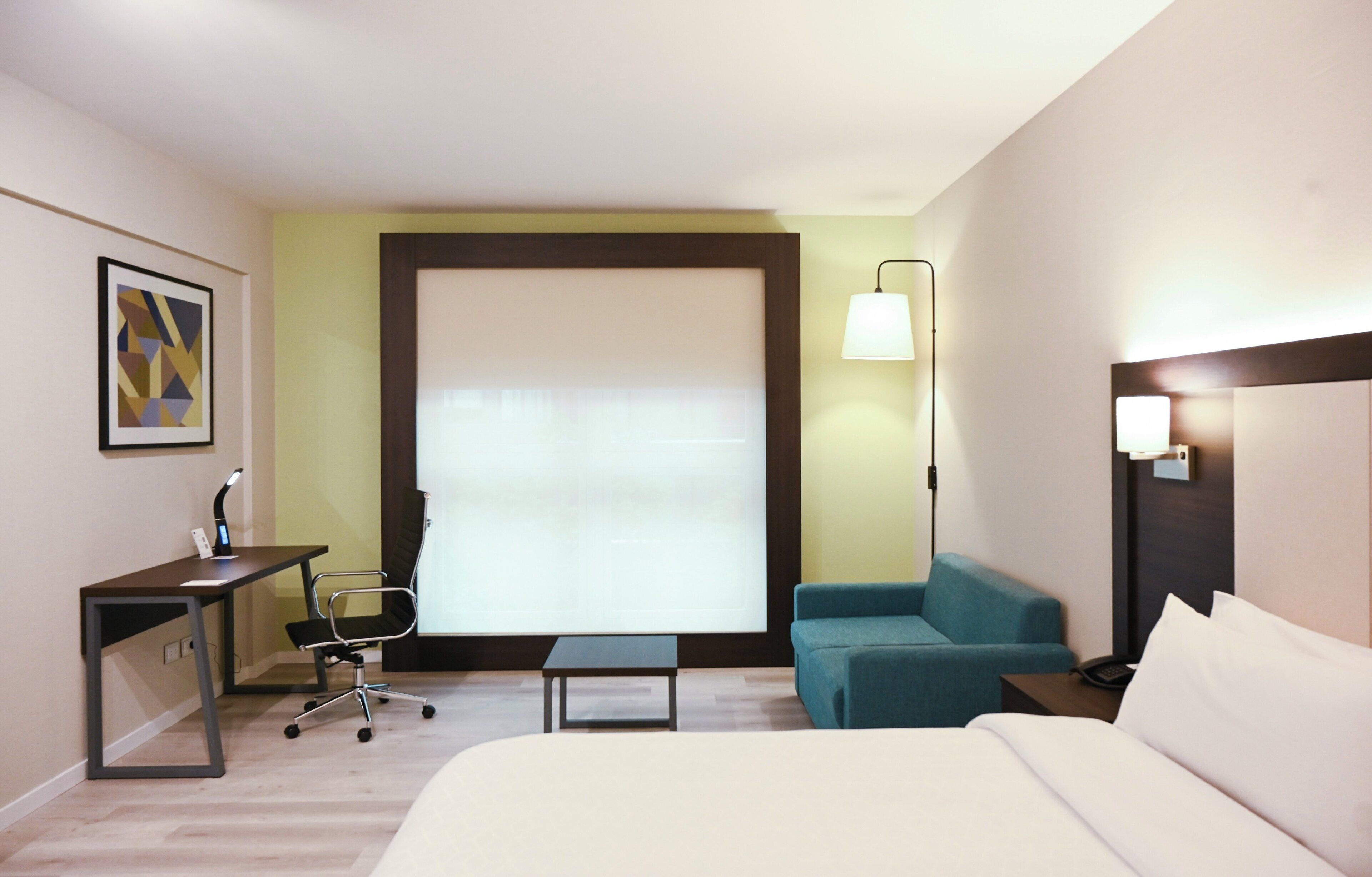 Habitación de hotel con cama, escritorio y silla, una mesita, sofá y ventana con persianas.