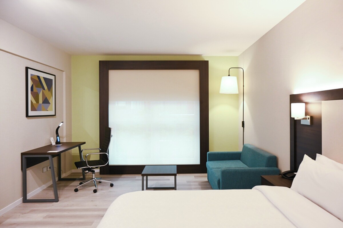 A hotel room with a bed, a desk with a chair, a small table, a sofa, and a window with blinds.