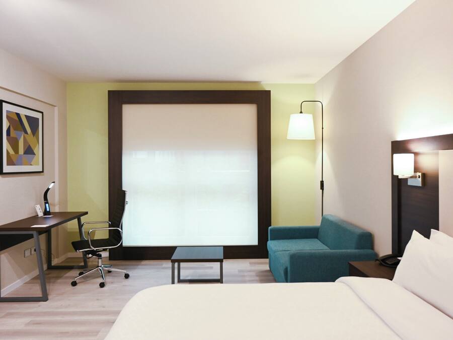 A hotel room with a bed, a desk with a chair, a small table, a sofa, and a window with blinds.