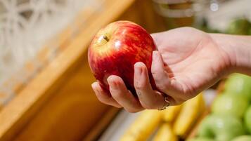 A hand holding a red apple, with a variety of fruits and vegetables in the background.