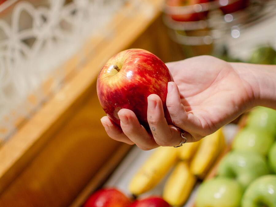 A hand holding a red apple, with a variety of fruits and vegetables in the background.