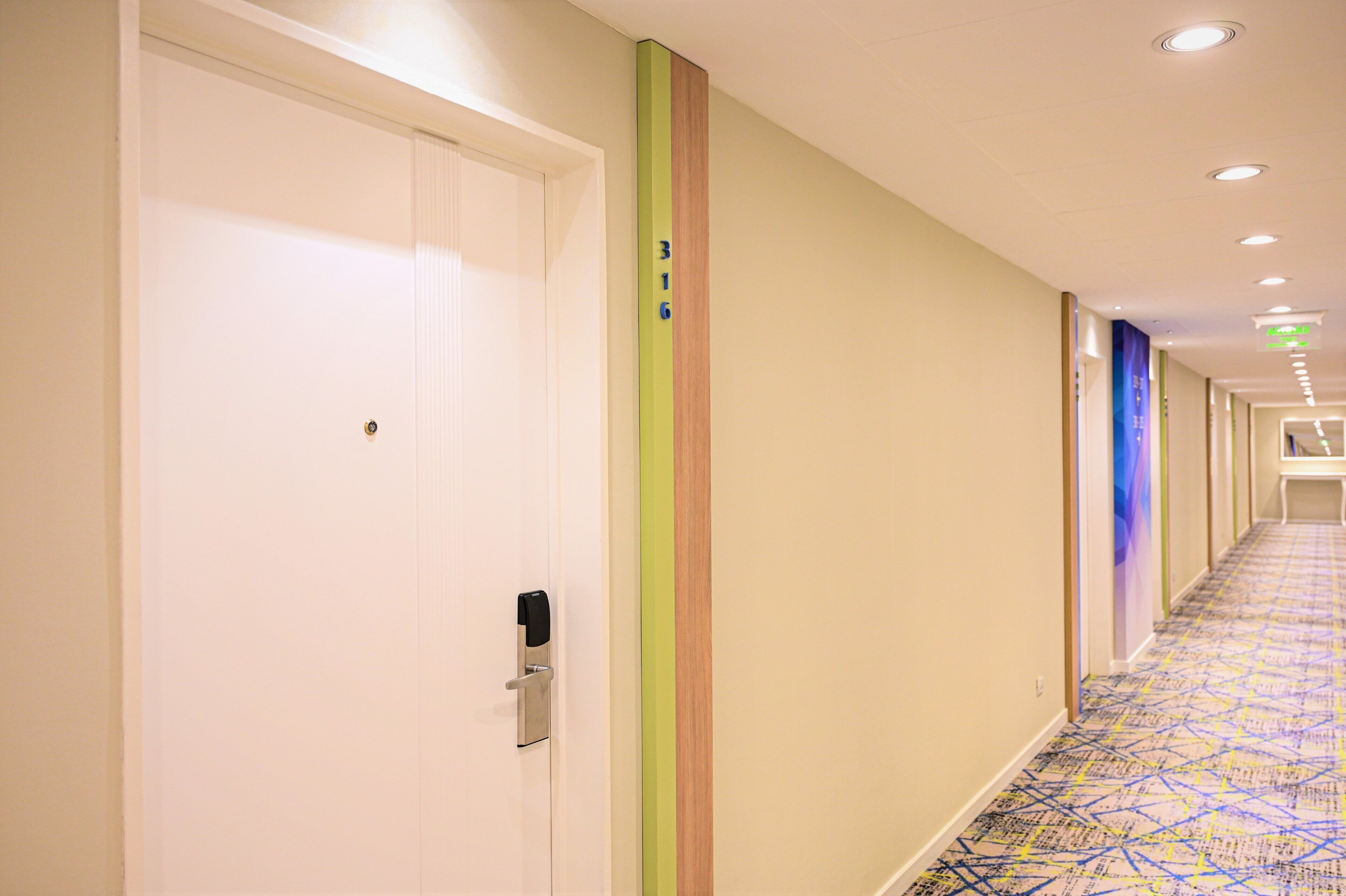 A hotel corridor with a patterned carpet, a white door with a keyhole, and a green vertical stripe on the wall.