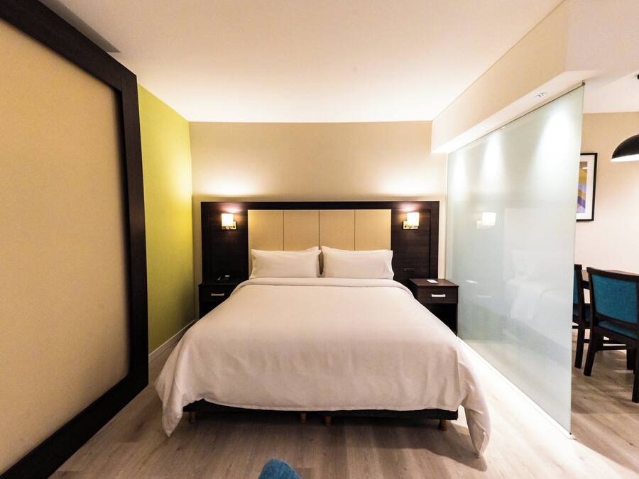 Una habitación de hotel con una cama grande, mesitas de noche, un escritorio con sillas y un separador de vidrio.