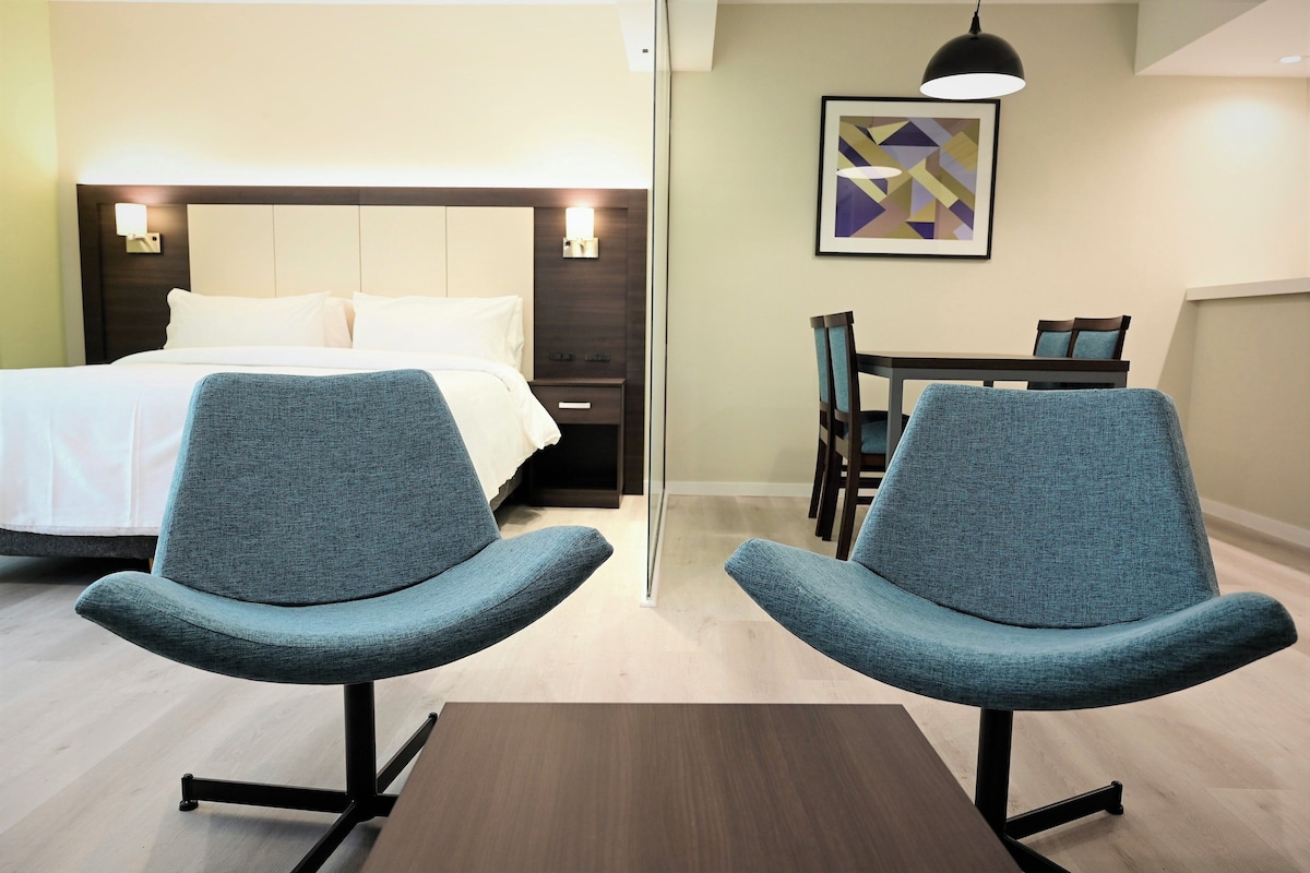 A hotel room with a bed, two chairs, a table, a desk, and a painting on the wall.