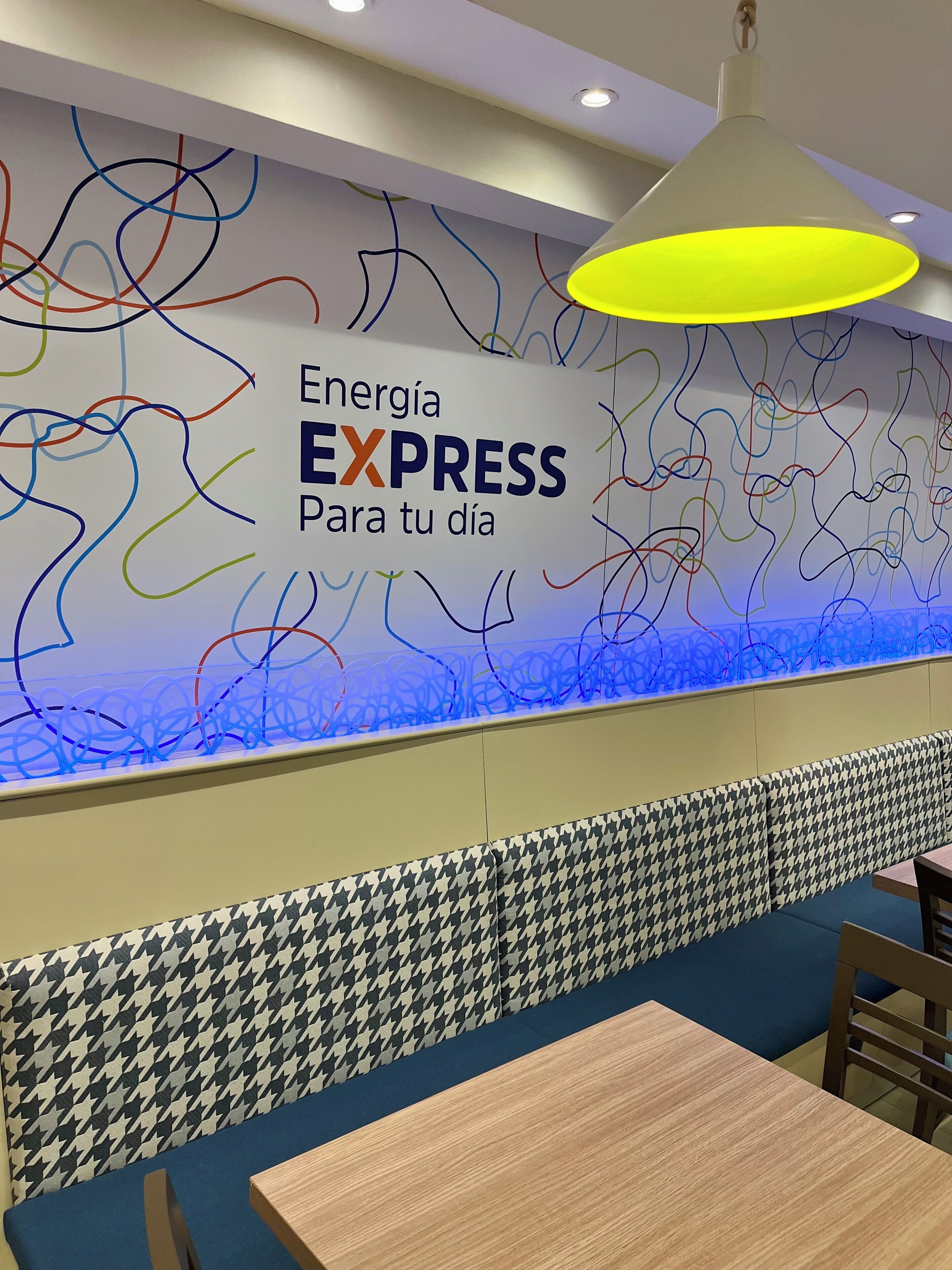 A restaurant interior with a wall featuring colorful abstract lines, a neon sign reading "Energía EXPRESS Para tu día", and a patterned houndstooth design on the lower wall.