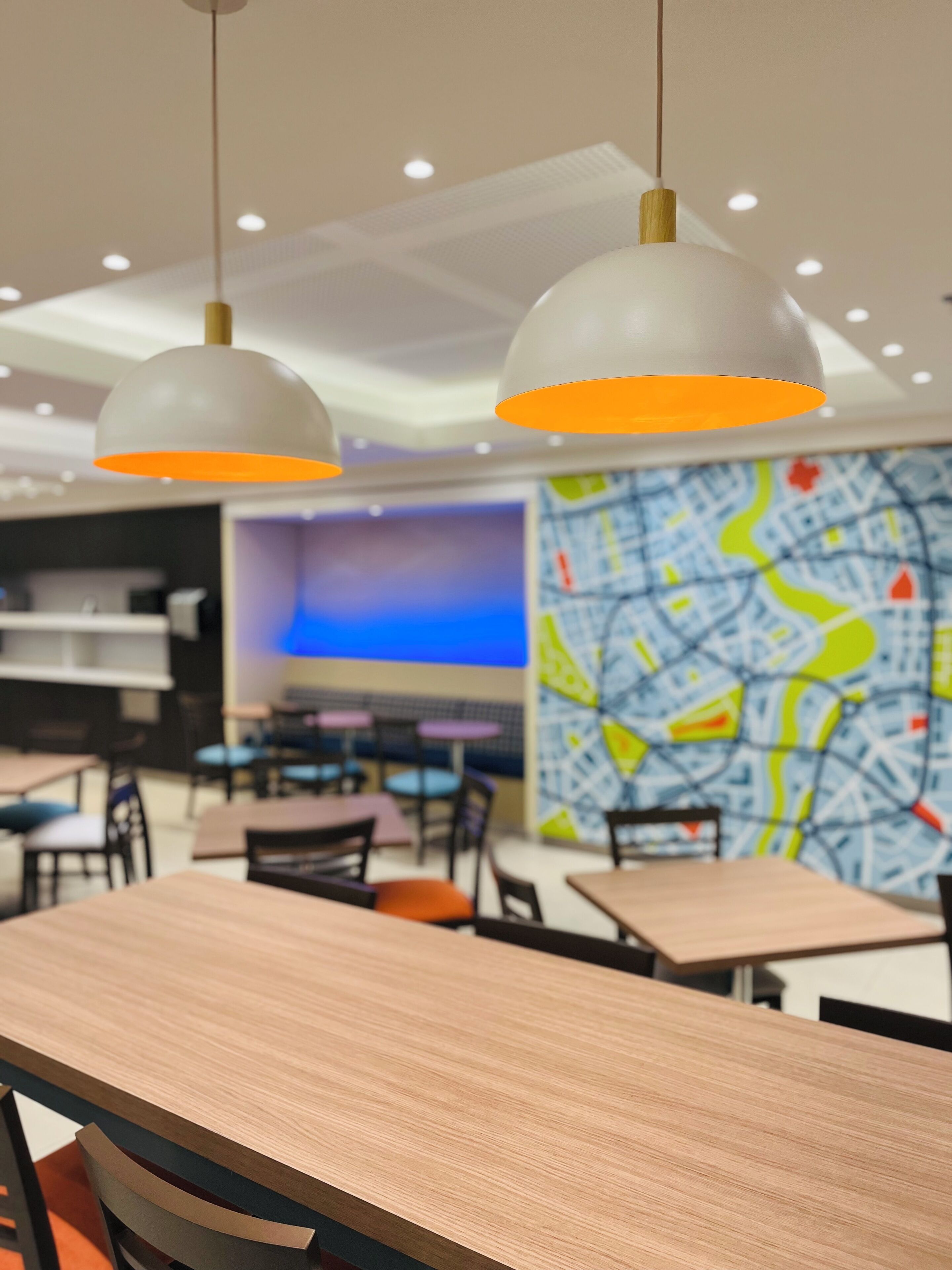Un espace de restauration moderne avec des tables et des chaises en bois, des suspensions dotées d’abat-jours orange et une grande fresque murale représentant une carte.