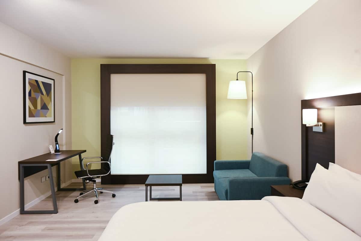 A hotel room with a bed, a desk with a chair, a small table, a sofa, and a window with blinds.