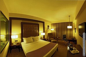 A hotel room with a large bed, two bedside tables with lamps, a desk with a chair, a TV, and a minibar. - Cambay Grand (Ahmedabad)