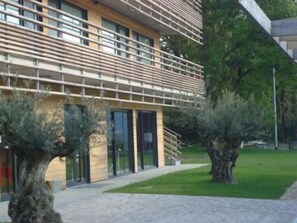 A modern building with a unique slatted facade, surrounded by well-maintained trees and a paved walkway. -  The Originals City, Hôtel Anaïade, Saint-Nazaire-Pornichet (Saint-Nazaire)