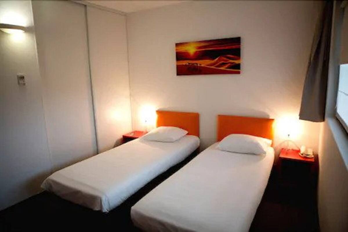 A hotel room with two single beds, each with a nightstand and a lamp. There is a painting on the wall above the beds.