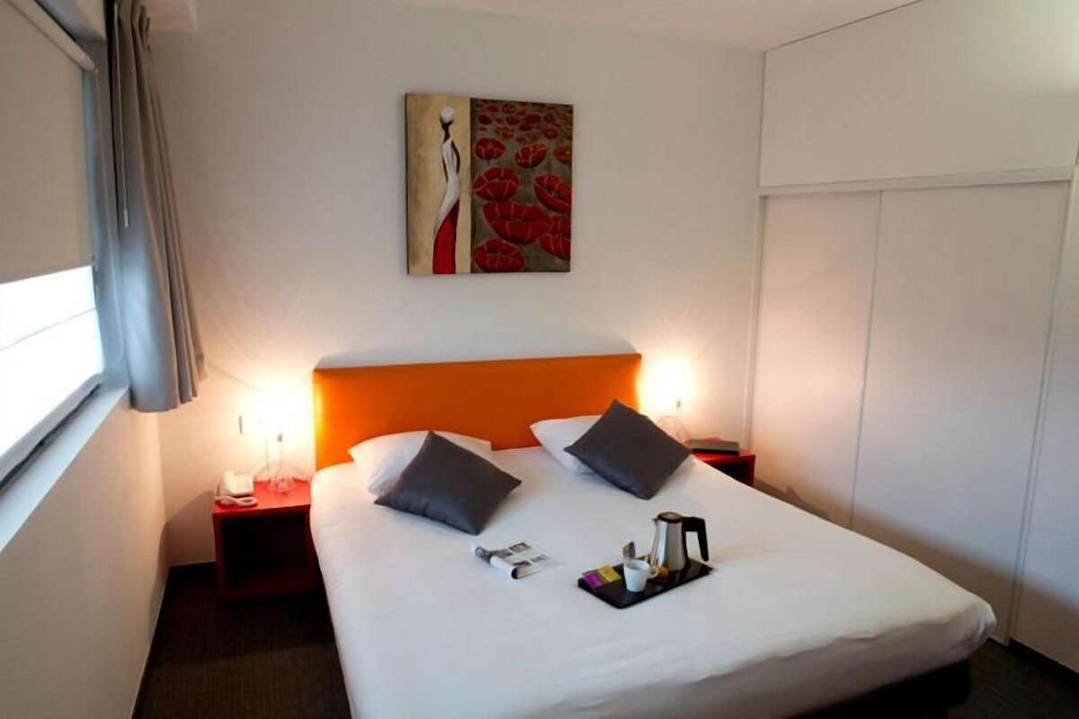 Habitación de hotel con una cama grande, dos almohadas, una mesita de noche con una lámpara, un cuadro en la pared y una bandeja con una taza y un libro.