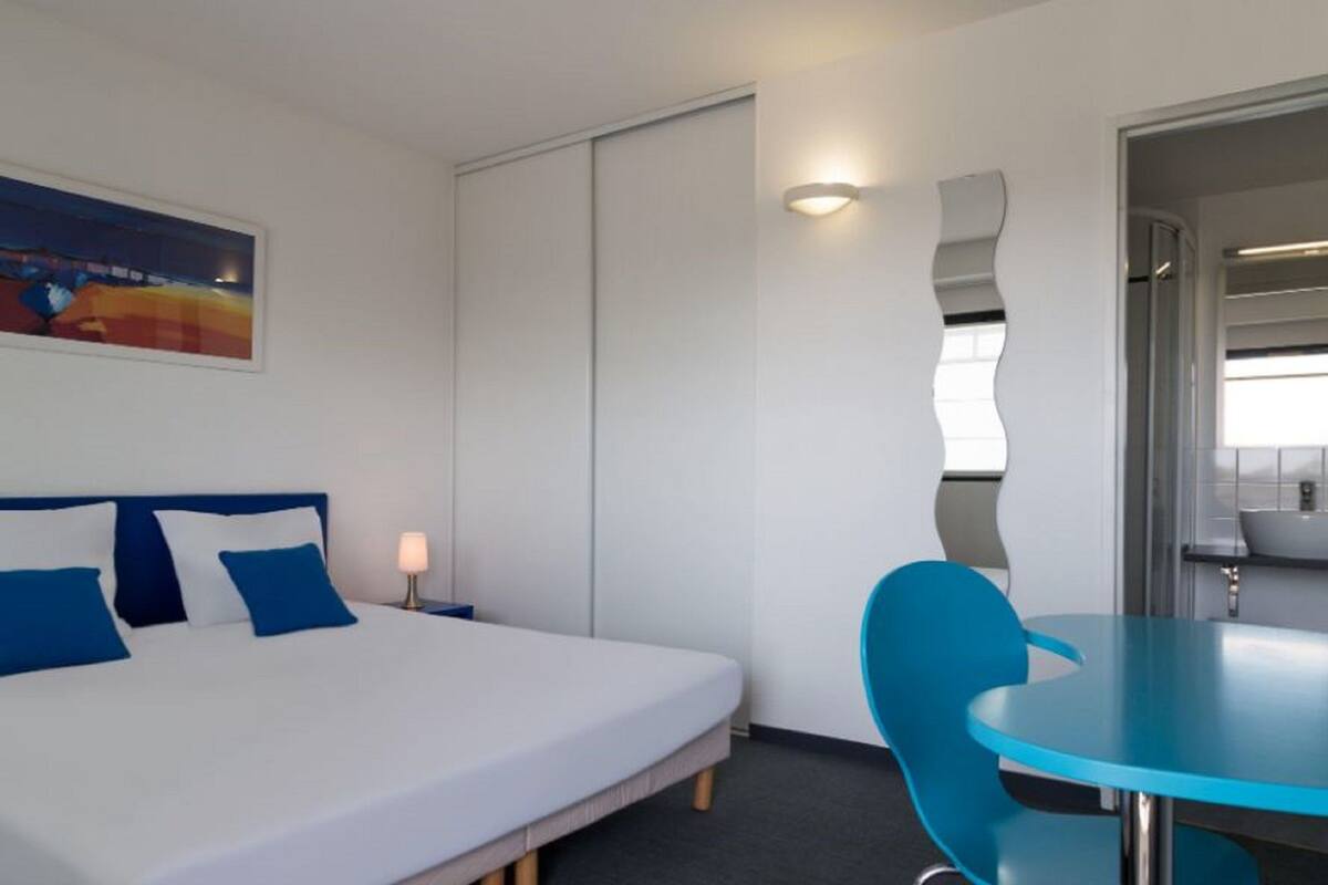 A hotel room with a bed, a blue chair, a small table, a nightstand, a lamp, a painting, and a bathroom visible through a door.