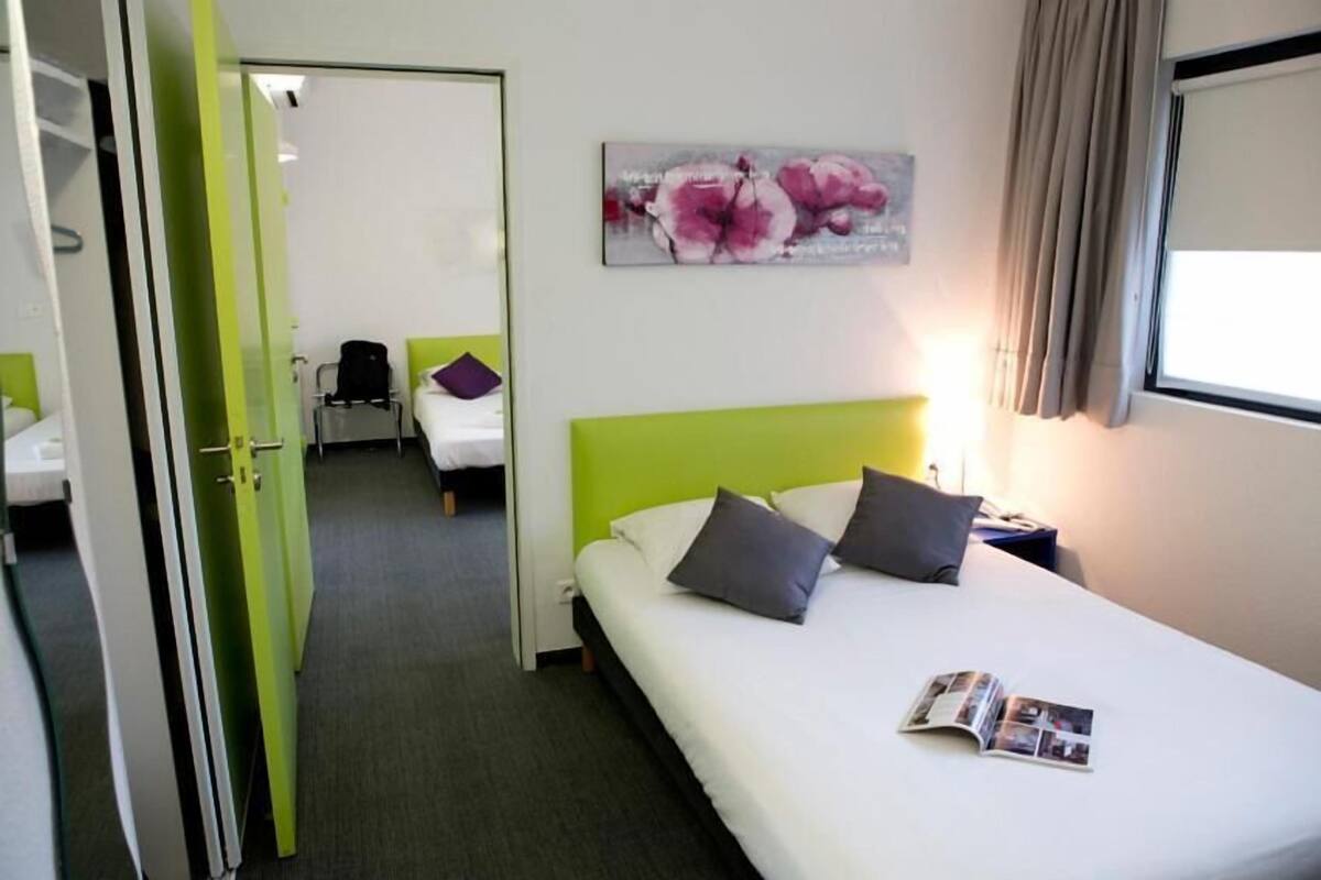 A hotel room with a green accent wall, a bed with two pillows, a nightstand with a lamp, and a painting of flowers on the wall.