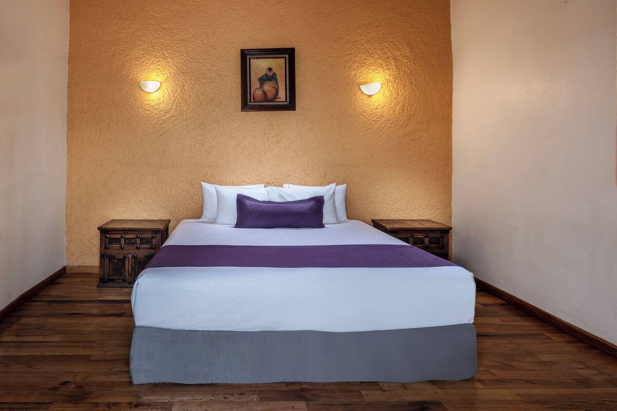A neatly made bed with a purple accent pillow, two bedside tables, and a framed picture on the wall.
